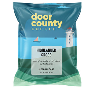 A bag of Door County Coffee Highlander Grogg medium roast coffee, featuring a blue and green coastal design with a lighthouse illustration. The image includes promotional text at the top and bottom on a white background.