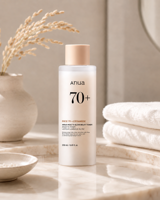 Anua 70+ Face 70 + Ceramide moisturizing toner bottle displayed on a marble surface with a neutral bathroom-style setting, including a vase with dried stems, folded white towels, and a mirror.