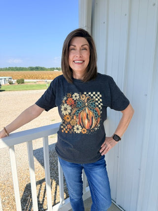 A woman stands on a porch wearing a dark charcoal heather graphic t-shirt with a fall harvest design featuring a pumpkin, flowers, and a checkered pattern, paired with blue jeans.
