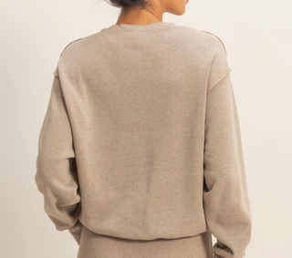 A person is shown from behind, wearing a plain crewneck sweatshirt in a light, warm beige or oatmeal tone, against a clean, off-white studio background.