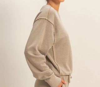 A side-profile view of a person wearing a light taupe-beige crewneck sweatshirt and matching drawstring pants against a plain off-white background.