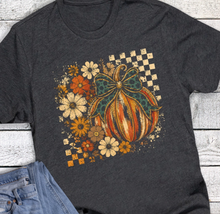 A dark charcoal gray t-shirt with a distressed autumn graphic featuring a striped pumpkin, a green bow, and wildflowers on a checkered background, styled flat-lay on a white wood plank surface with blue jeans partially visible at the bottom left.